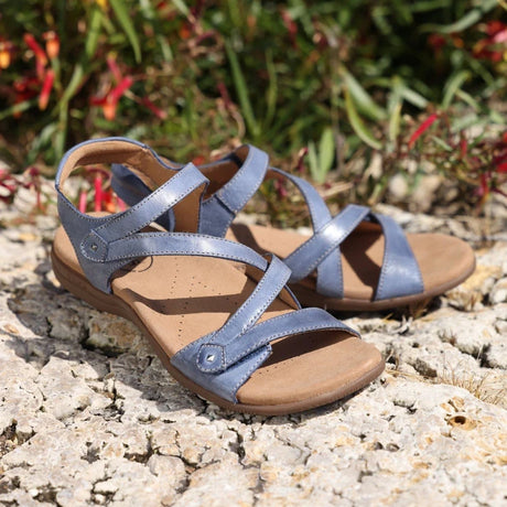 Pair of blue Taos sandals with brown soles on a stone surface with greenery in the background