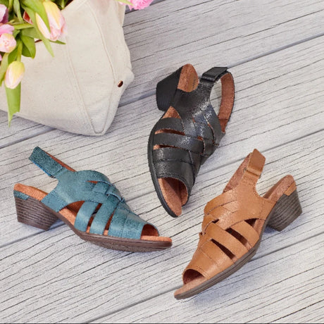 Three pairs of Taos sandals in blue, black, and brown on a wooden surface with flowers and a bag in the background.