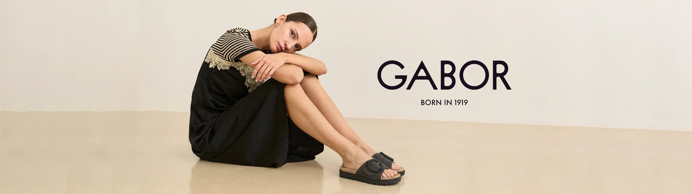 Woman sitting on a beige floor wearing black sandals with 'Gabor' branding.